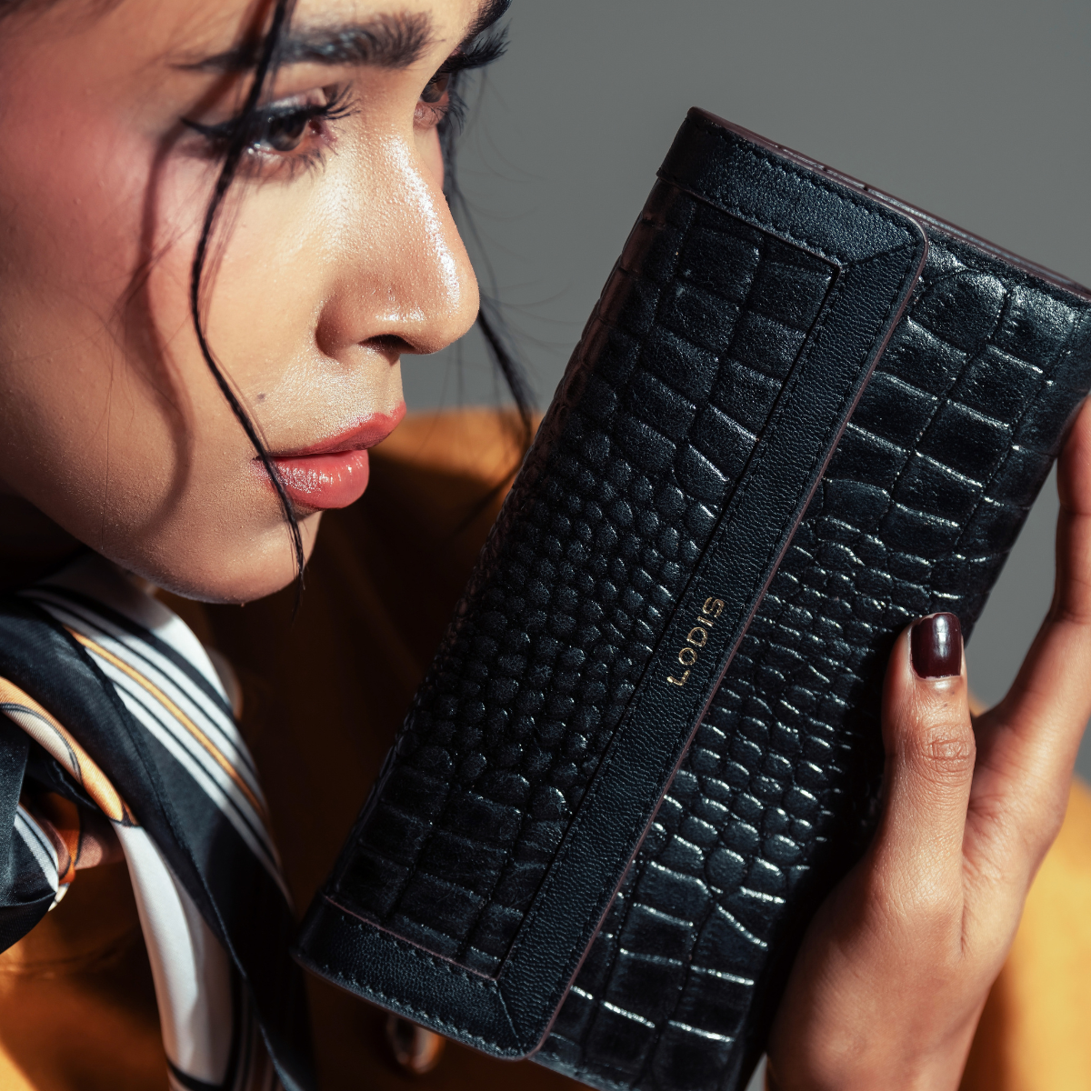 Woman Holding Lodis Amara Checkbook Clutch - Fall Lifestyle Shot with Striped Silk Scarf and Mustard Top in Studio Setting, Glossy Croco-Embossed Leather with Gold Foil Lodis Logo | Lodis 1965