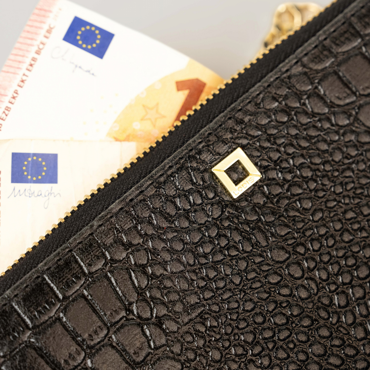 Lodis Amara Sling in Black - Close-Up of Top Zip Opening and Gold-Tone Square Logo Plate on Black Glossy Croco-Embossed Leather, Shown with Banknotes Inside | Lodis 1965