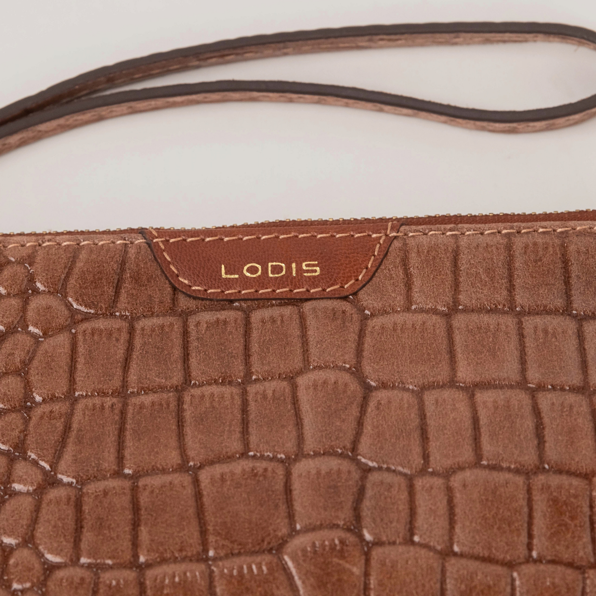 Lodis Amara Zip Pouch in Chestnut - Close-Up of Gold Foil Lodis Logo on Smooth Texas Leather Tab with Wristlet Strap Above Croco-Embossed Texture | Lodis 1965
