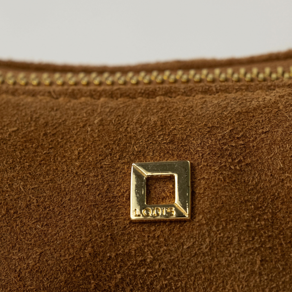 Lodis Axis Crossbody in Tan - Close-Up of Gold-Tone Square Logo Plate and Gold-Tone Zip Edge on Golden Tan Suede | Lodis 1965