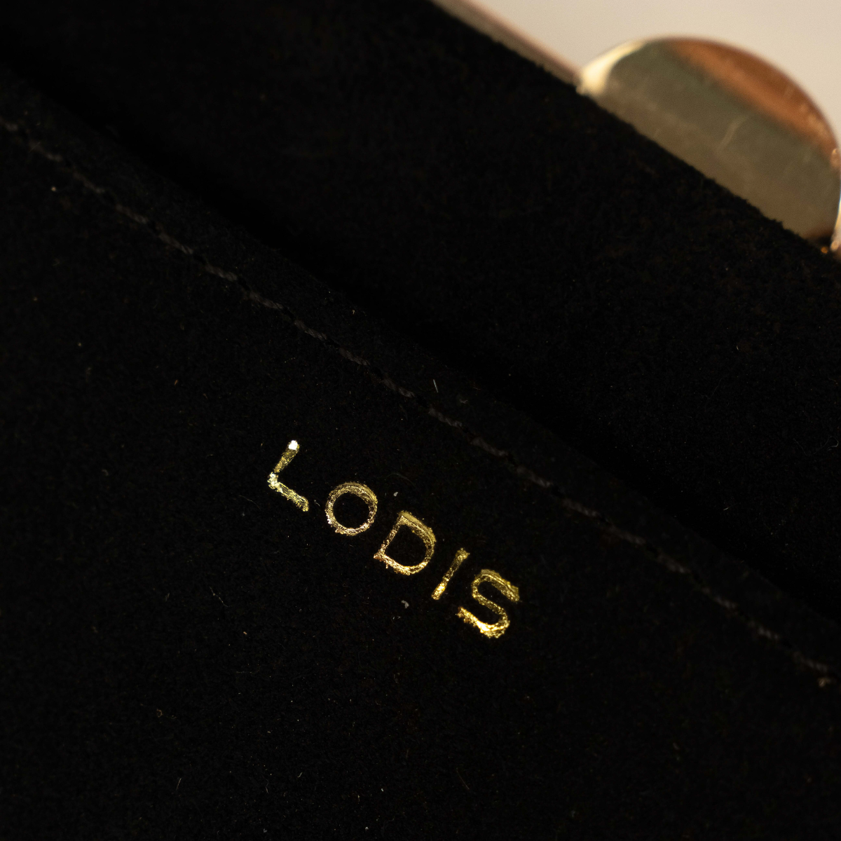 Lodis Celara Frame Clutch in Black - Macro Close-Up of Gold Foil Lodis Logo on Black Texas Suede Pocket with Gold-Tone Clasp in Background | Lodis 1965