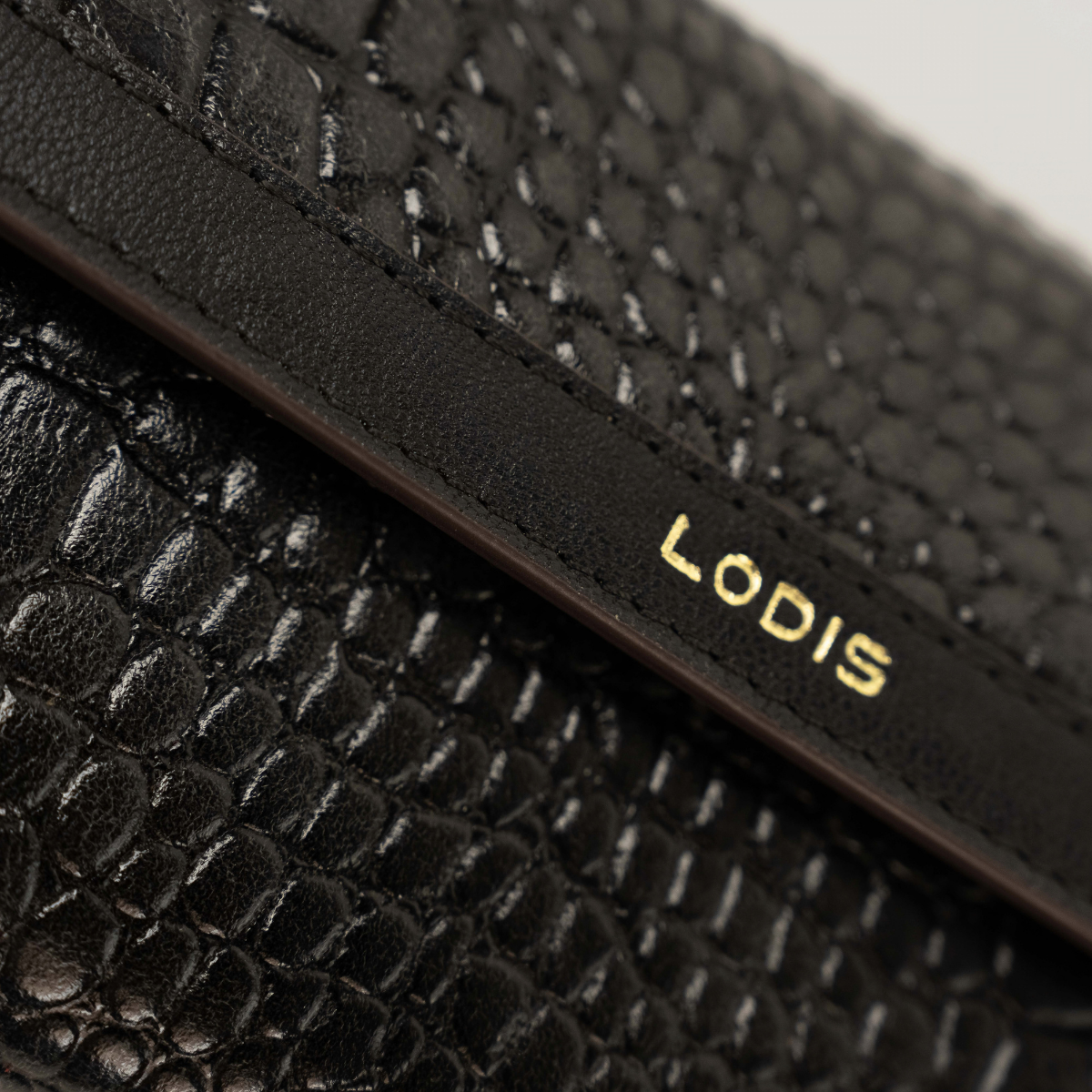 Lodis Amara Checkbook Clutch in Black - Close-Up of Gold Foil Lodis Logo on Stitched Texas Leather Band Across Black Glossy Croco-Embossed Flap | Lodis 1965