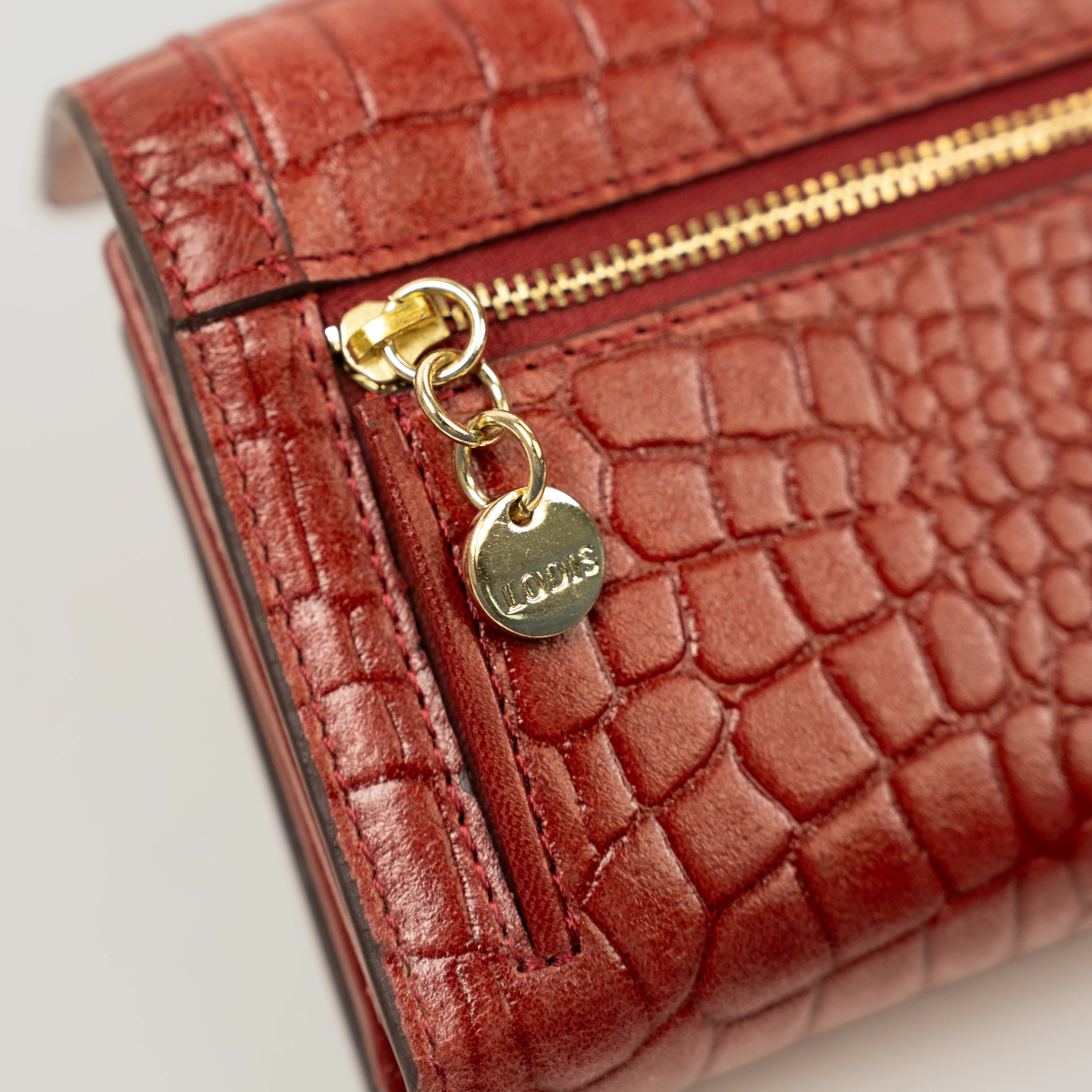 Lodis Amara Checkbook Clutch in Samba - Close-Up of Gold-Tone Zipper Pull with Engraved Lodis Coin Charm on Back Zip Pocket, Deep Red Glossy Croco-Embossed Leather | Lodis 1965