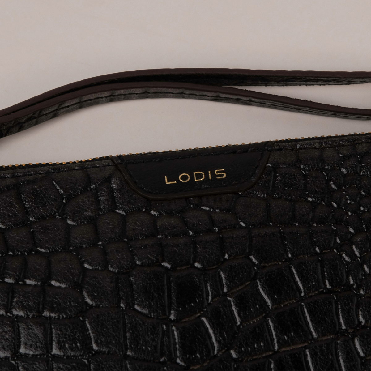 Lodis Amara Zip Pouch in Black - Close-Up of Gold Foil Lodis Logo on Smooth Texas Leather Tab with Wristlet Strap Above Croco-Embossed Texture | Lodis 1965