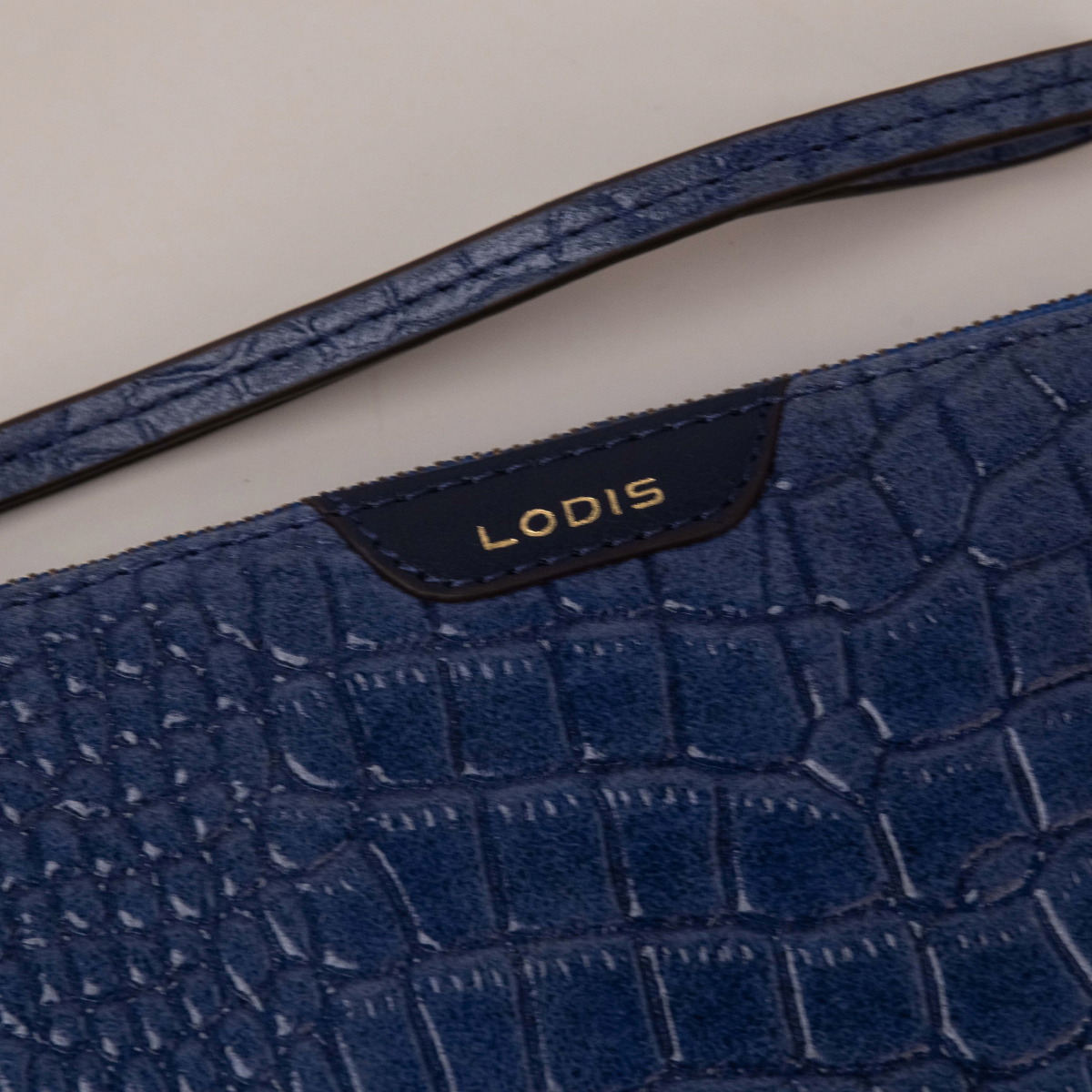 Lodis Amara Zip Pouch in Cobalt - Close-Up of Gold Foil Lodis Logo on Smooth Texas Leather Tab with Wristlet Strap Above Croco-Embossed Texture | Lodis 1965