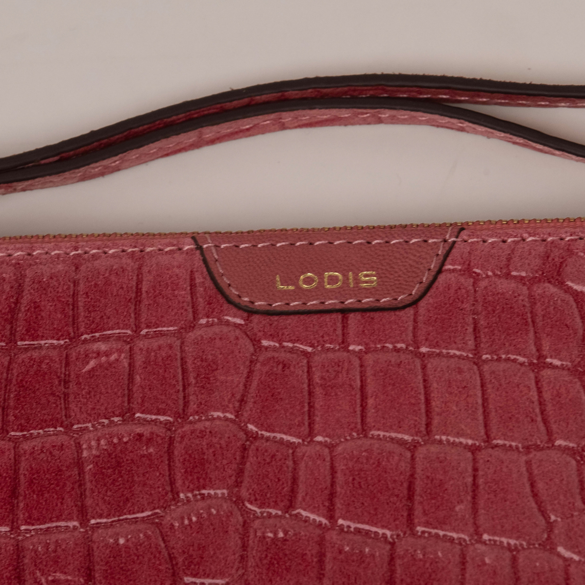 Lodis Amara Zip Pouch in Glossy Pink - Close-Up of Gold Foil Lodis Logo on Smooth Texas Leather Tab with Wristlet Strap Above Croco-Embossed Texture | Lodis 1965