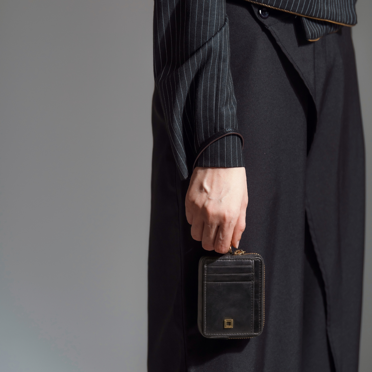 Woman Carrying Lodis Blair Trouble Card Case in Black at Her Side - Fall Lifestyle Shot with Black Pinstripe Blazer and Wide-Leg Black Trousers in Studio Setting, Glazed Leather | Lodis 1965